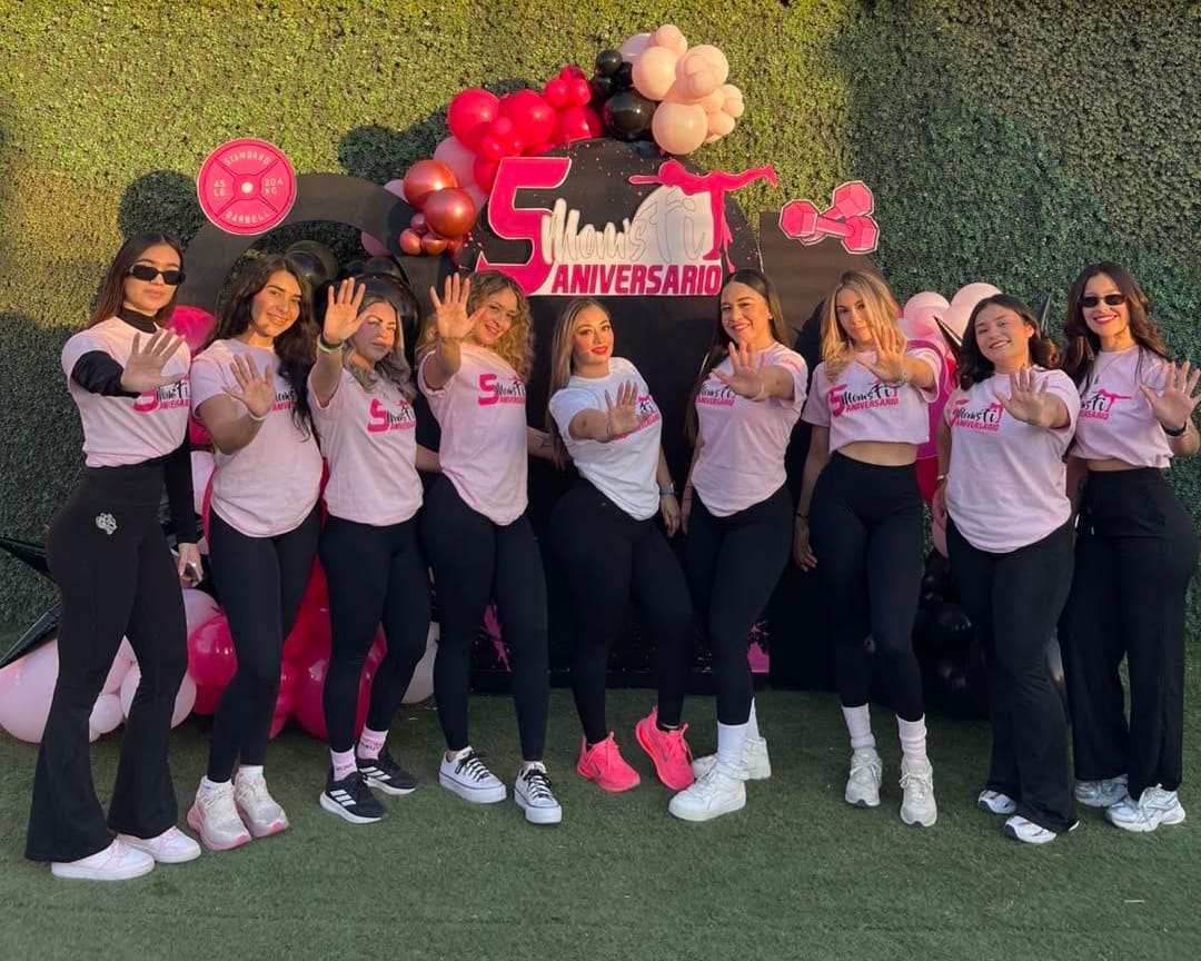 Alumnas de rosa con la playera del quinto aniversario, saludando frente al arco de globos