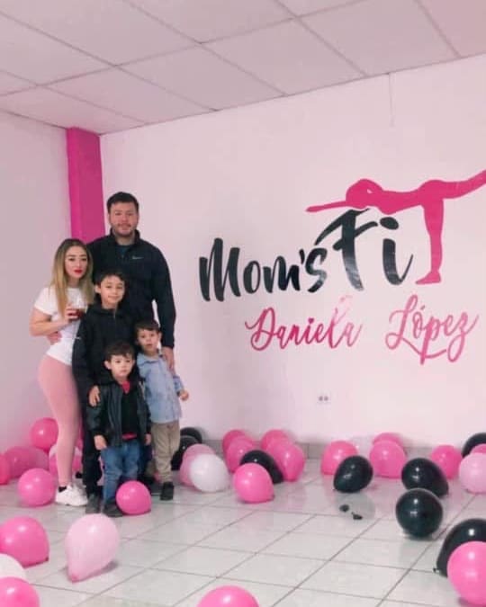 Una familia entre globos rosas y negros, frente al logotipo de Moms Fit pintado en la pared
